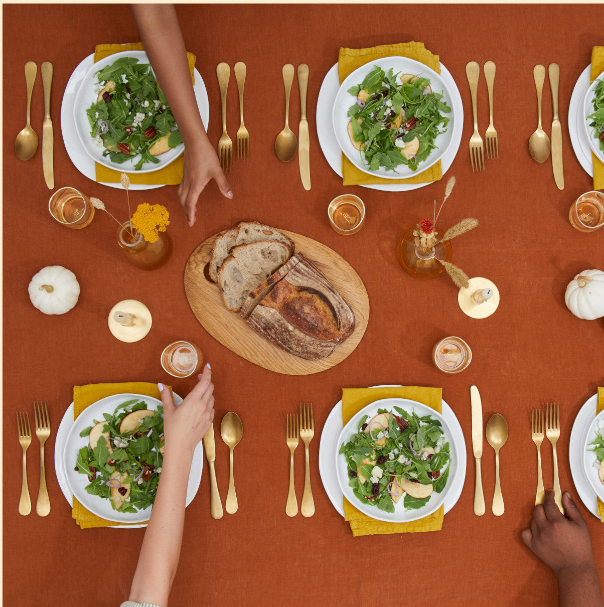 A dining table set with orange tablecloth, gold utensils, and plates of salad. A wooden board with sliced bread is in the center. People's hands are reaching for the bread. Small pumpkins are on the table.
