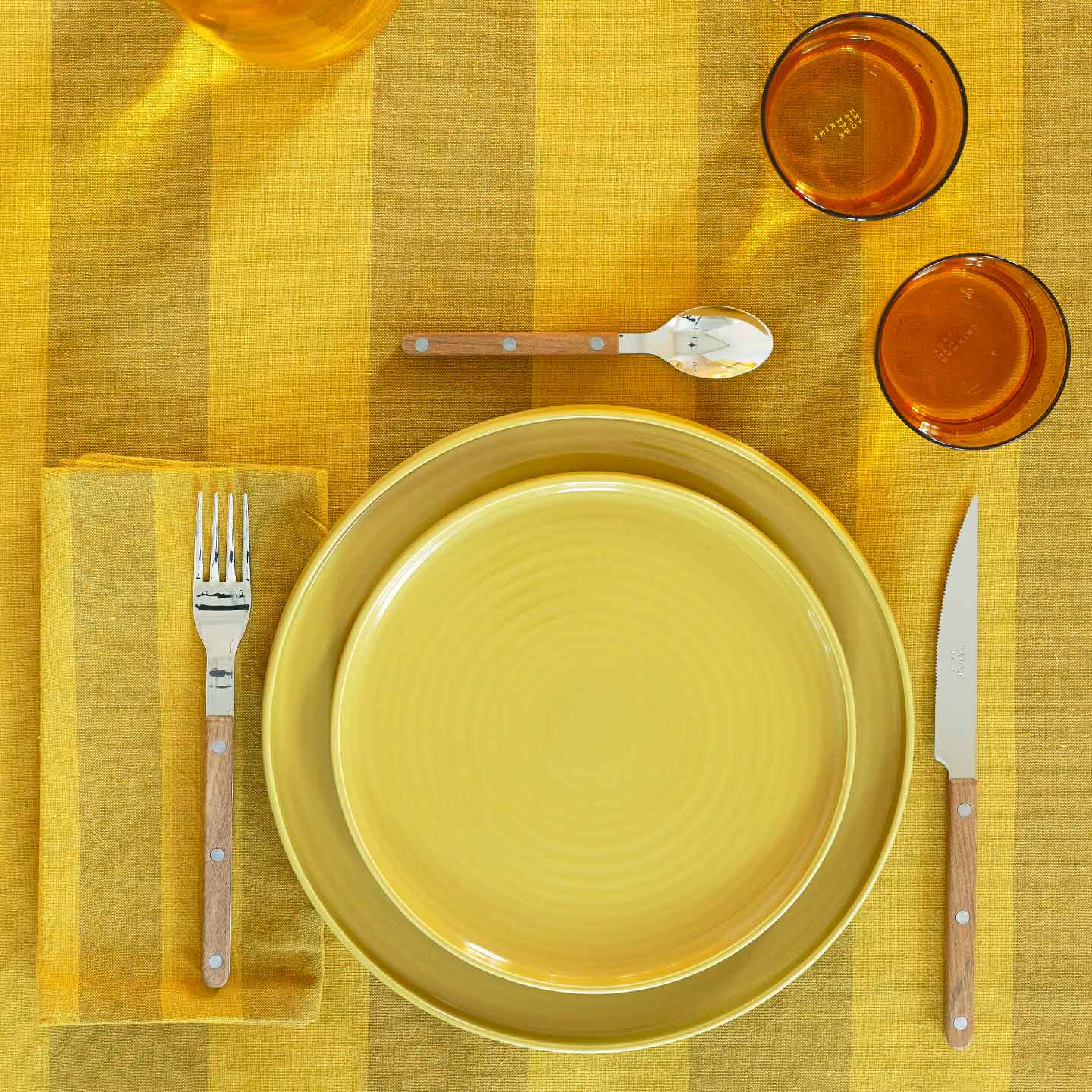 Placesetting with Essential Striped Dinner Napkin in Mustard and Bronze on yellow striped tablecloth.