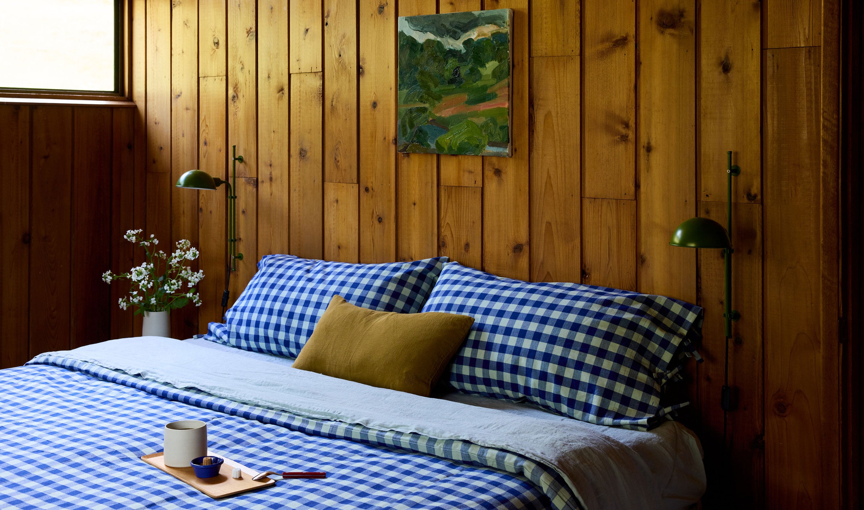 A cozy bedroom scene with a bed dressed in blue and white gingham bedding, a mustard yellow accent pillow, and a small landscape painting hanging on a warm wood-paneled wall. Two green wall lamps, a vase of flowers, and a tray with a mug sit beside the bed.