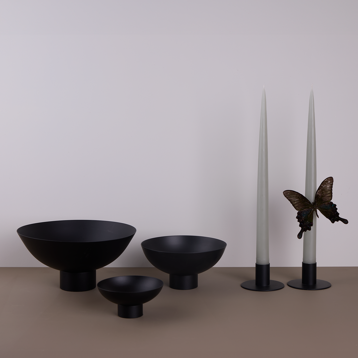 A trio of footed bowls and pair of candle holders, all in Black, with grey taper candles. Styled with a butterfly.