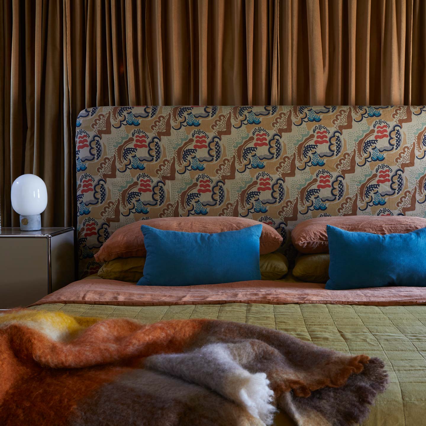 Bed with patterned headboard and bedding in terracotta, peacock, bronze, and multicolor throw.