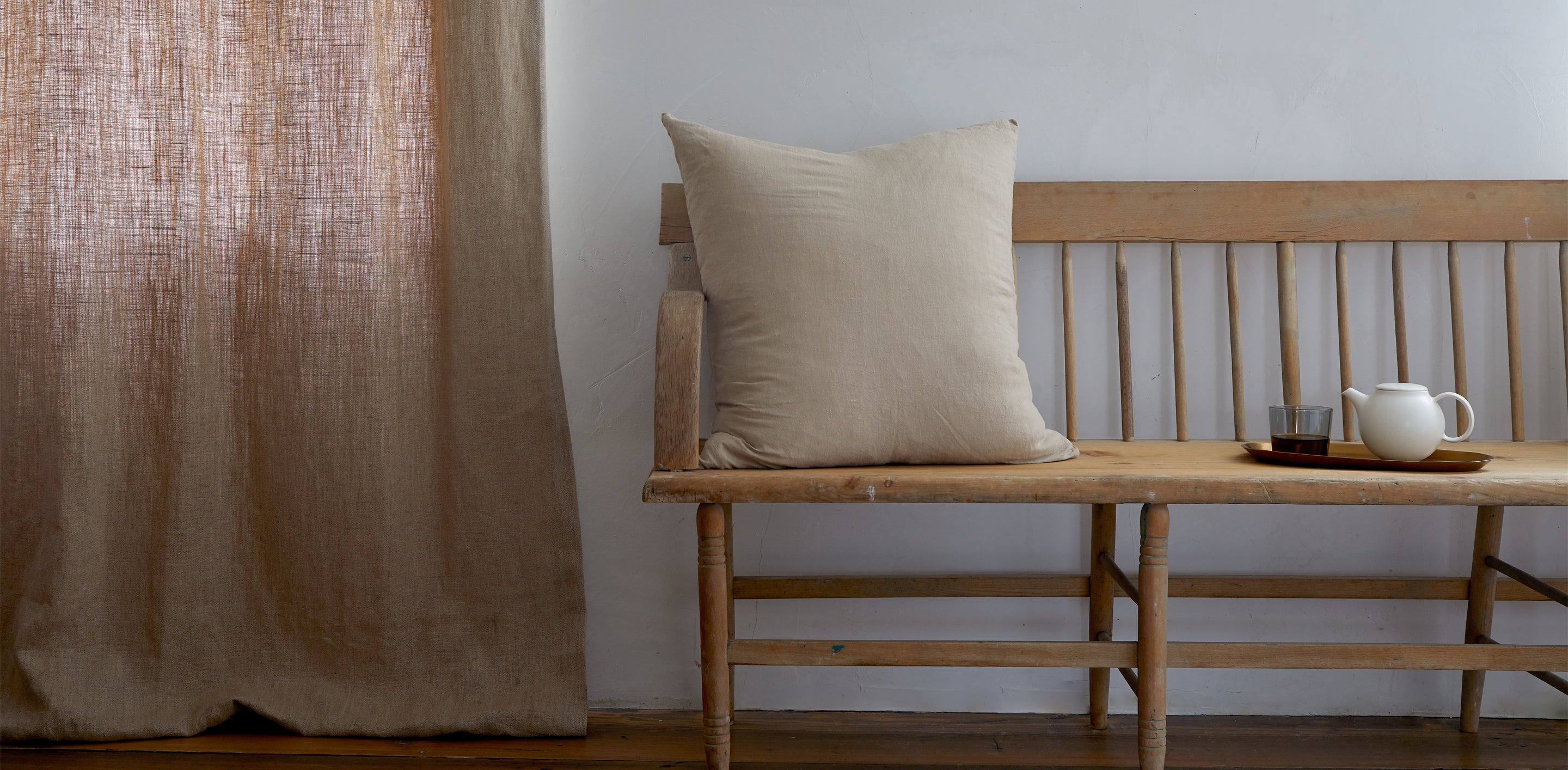 A wooden bench against a white wall has a large beige cushion. A tray with a teapot and a small cup sits on the bench. Beige curtains hang to the left.