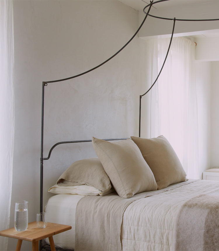 A minimalist bedroom features a metal canopy bed with beige bedding, two pillows, and a wooden side table with a glass of water and a carafe. Soft, natural light filters through sheer curtains.