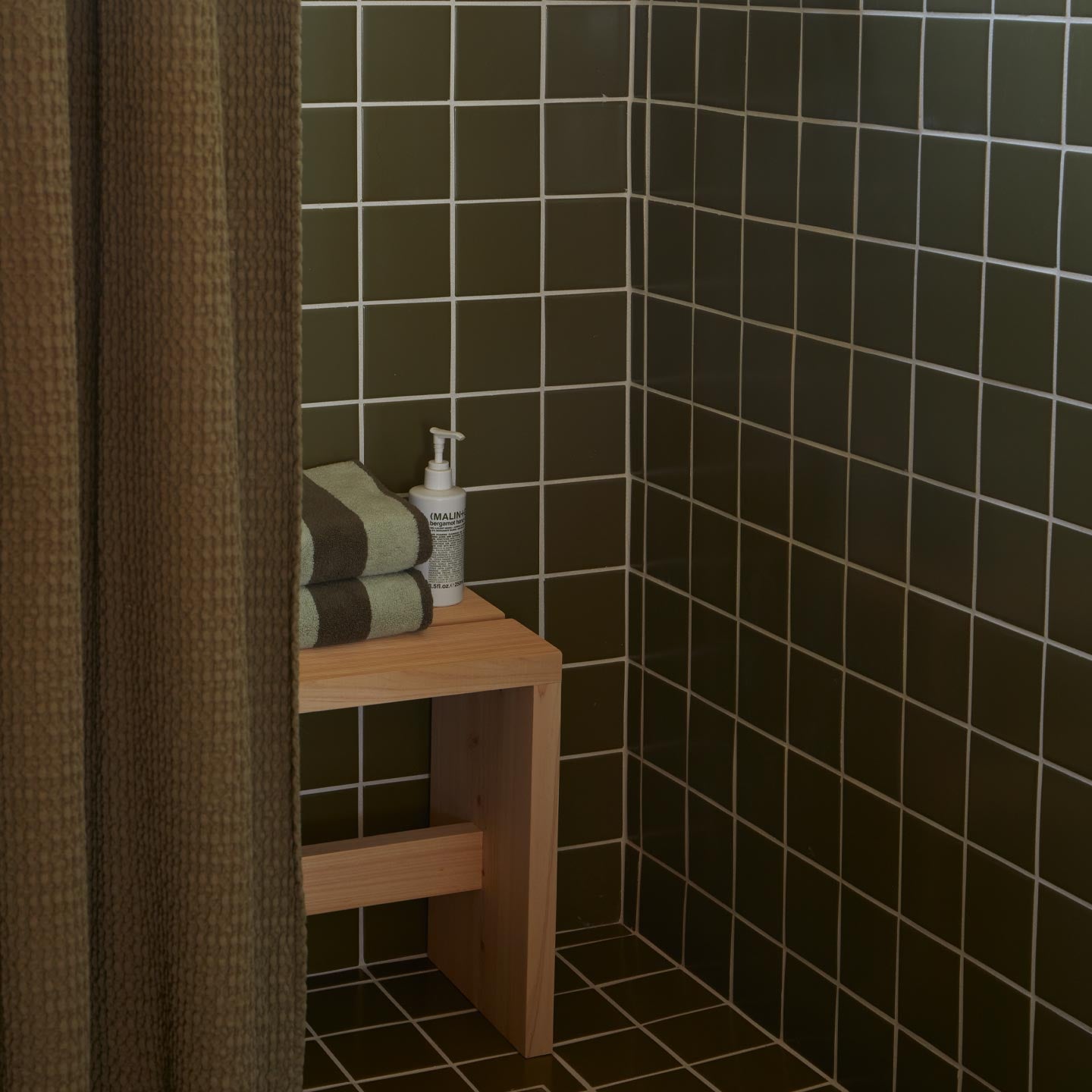 Tiled shower with olive shower curtain and wooden bench with towels and soap.