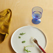 Overhead of place setting on wood tabletop with essential Gingham Napkin in Mustard, Blue Seeded Glass, and Simple Porcelain Plate with red fork.