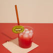 Red cocktail with ice and a kiwi slice on a blush cocktail napkin on a red surface.