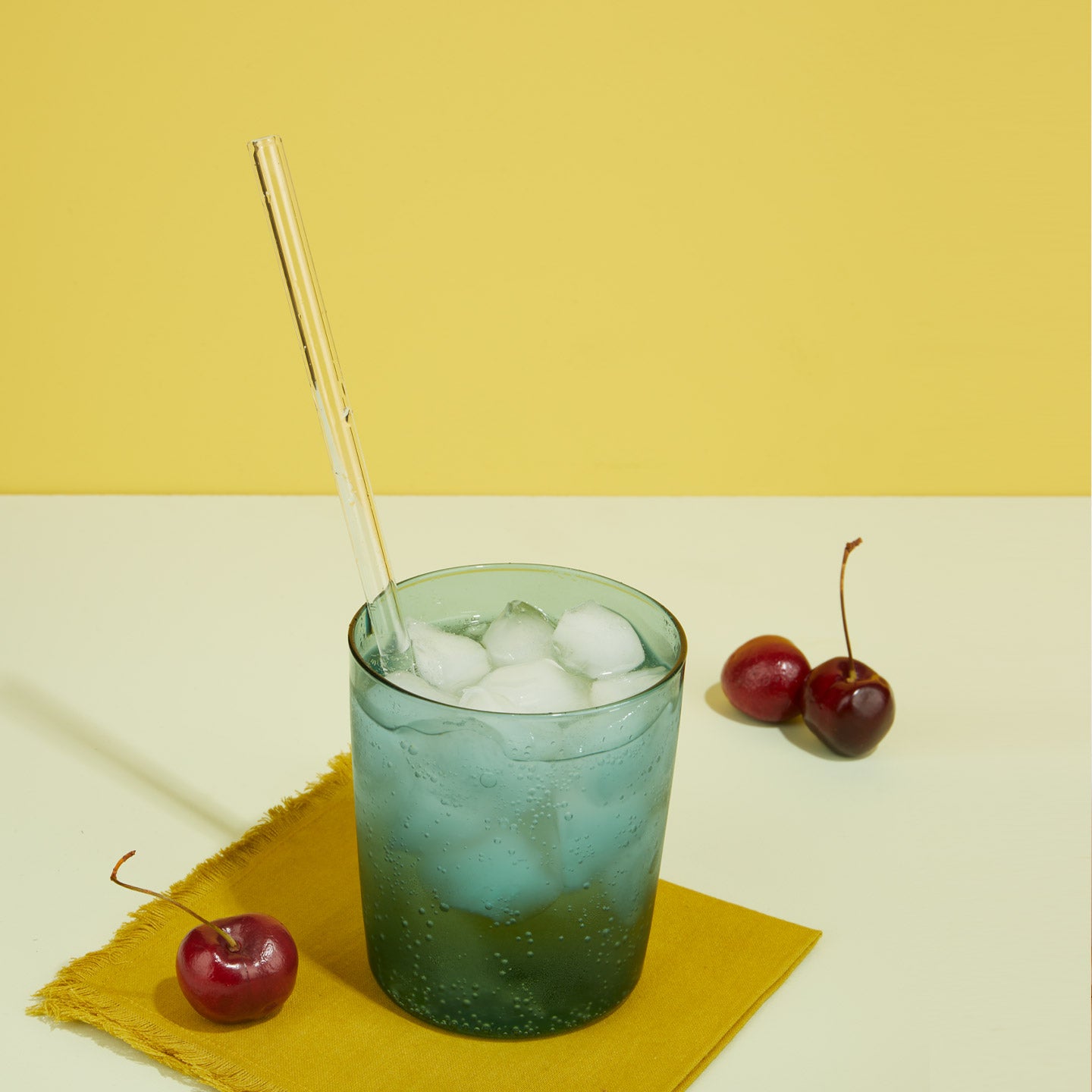 Essential Glass Straw in Essential Peacock Glass with ice and cocktail napkin and cherries.