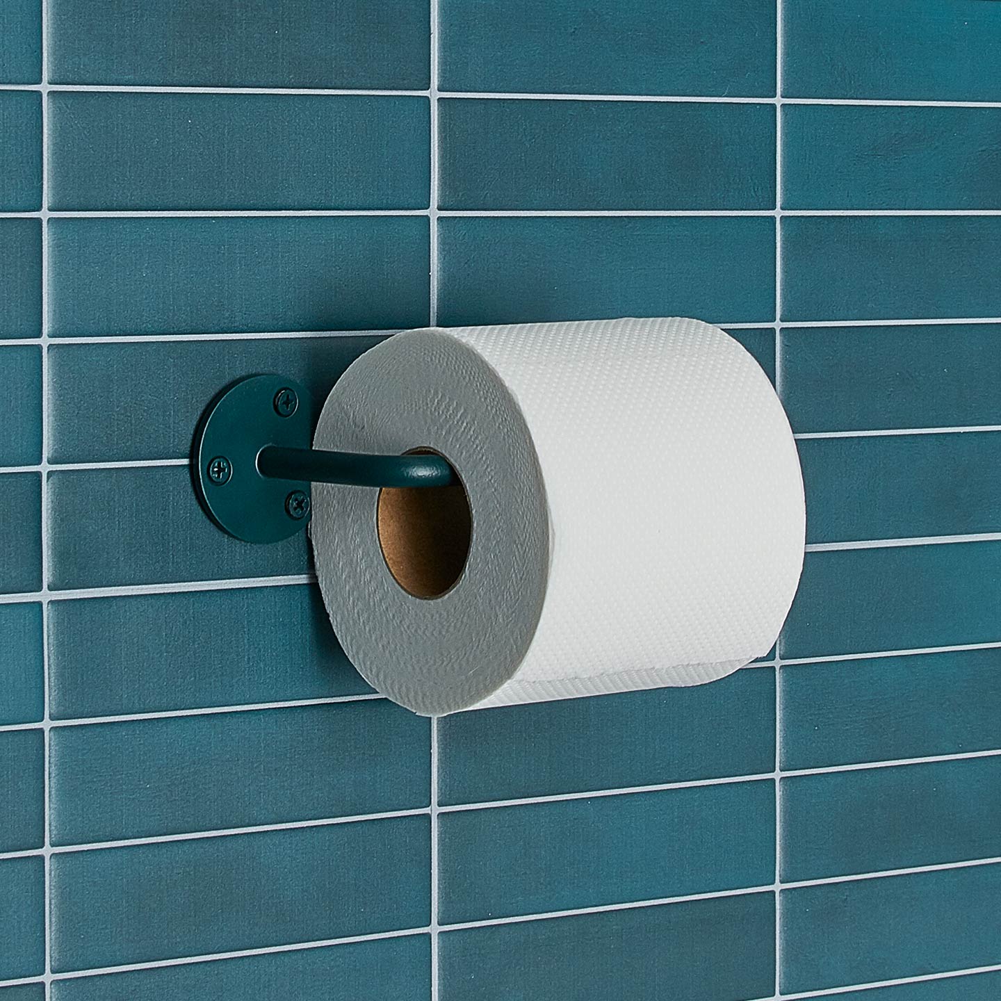 Essential toilet paper holder in Dark Peacock with a roll of toilet paper on tiled wall.