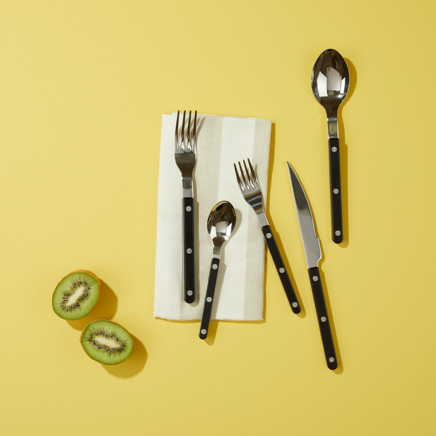 Essential Striped Napkin in Ivory with black flatware and kiwi on yellow background.