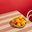 Oranges in Mustard Essential Low Bowl on Striped blush Tablecloth with chair and red background.