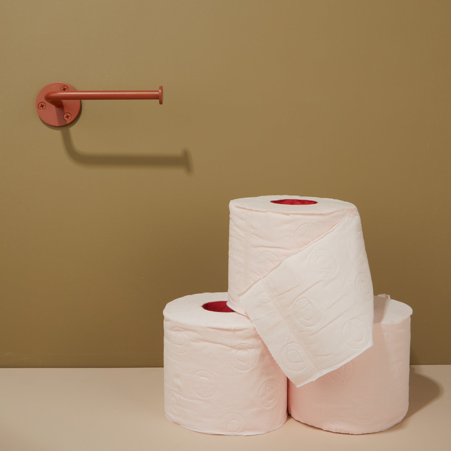 Three rolls of toilet paper stacked on a beige surface with a brown wall and terracotta toilet paper holder in the background.