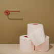 Three rolls of toilet paper stacked on a beige surface with a brown wall and terracotta toilet paper holder in the background.