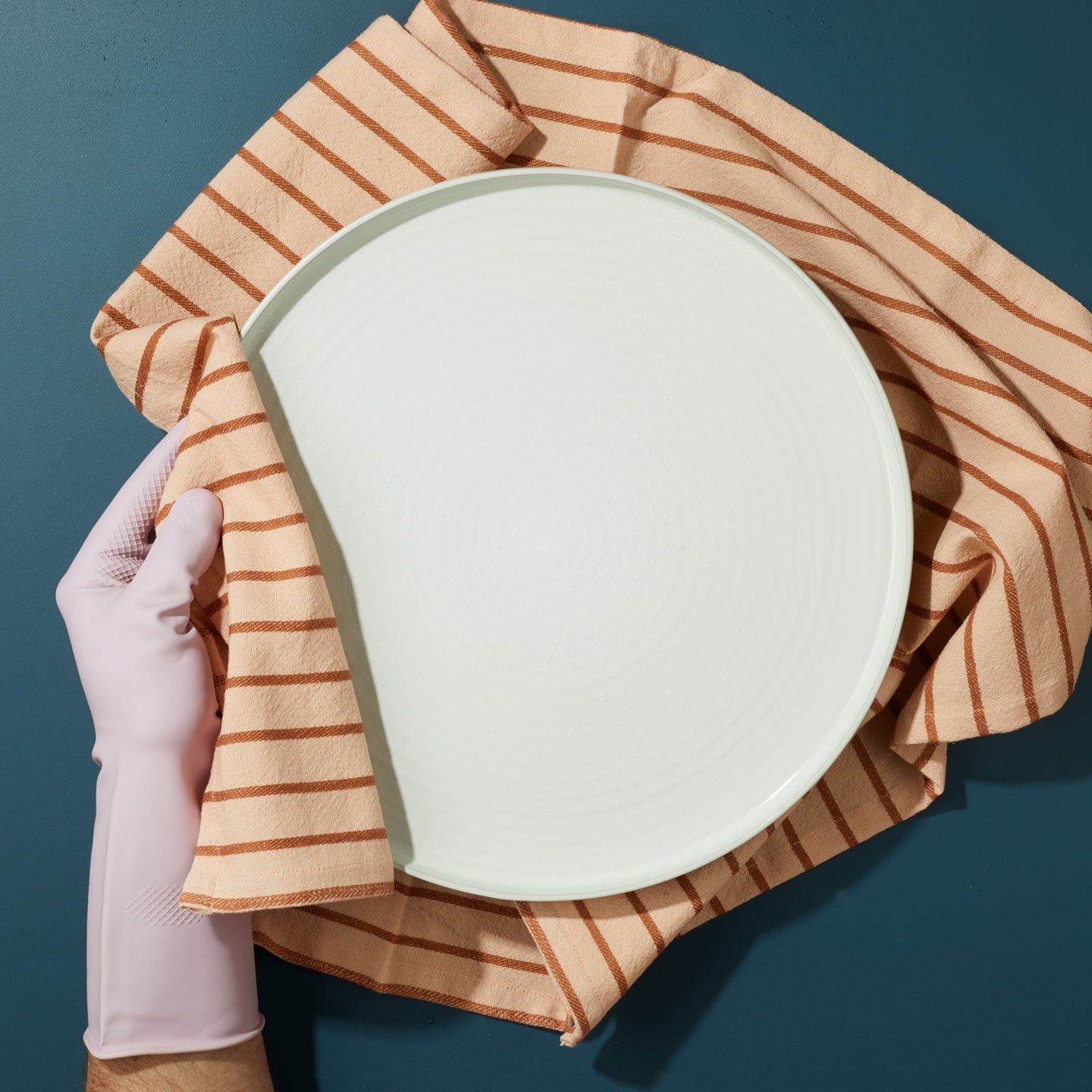 White plate held by a gloved hand on an Essential striped towel in blush against a blue background