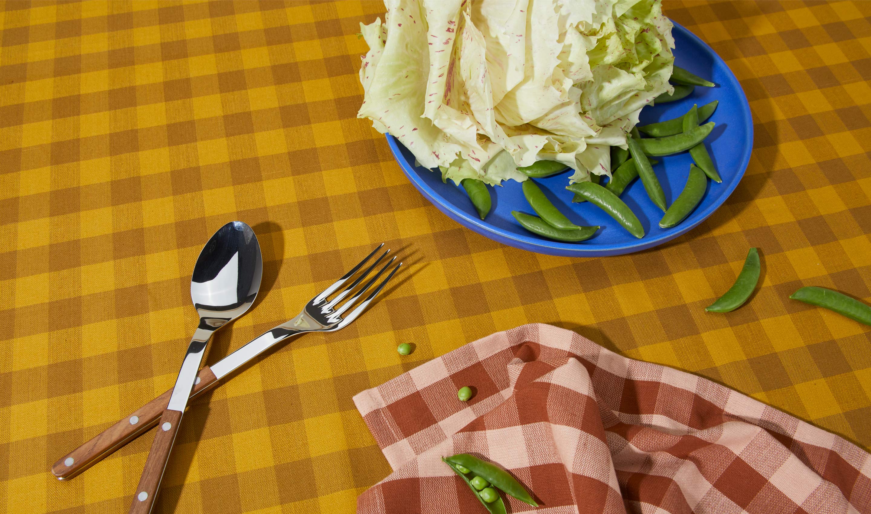 A tabletop covered in a mustard/bronze gingham tablecloth. The table is arranged with a blue plate holding lettuce and green beans. Nearby a teak-handled spoon and fork and a blush/terracotta gingham napkin.