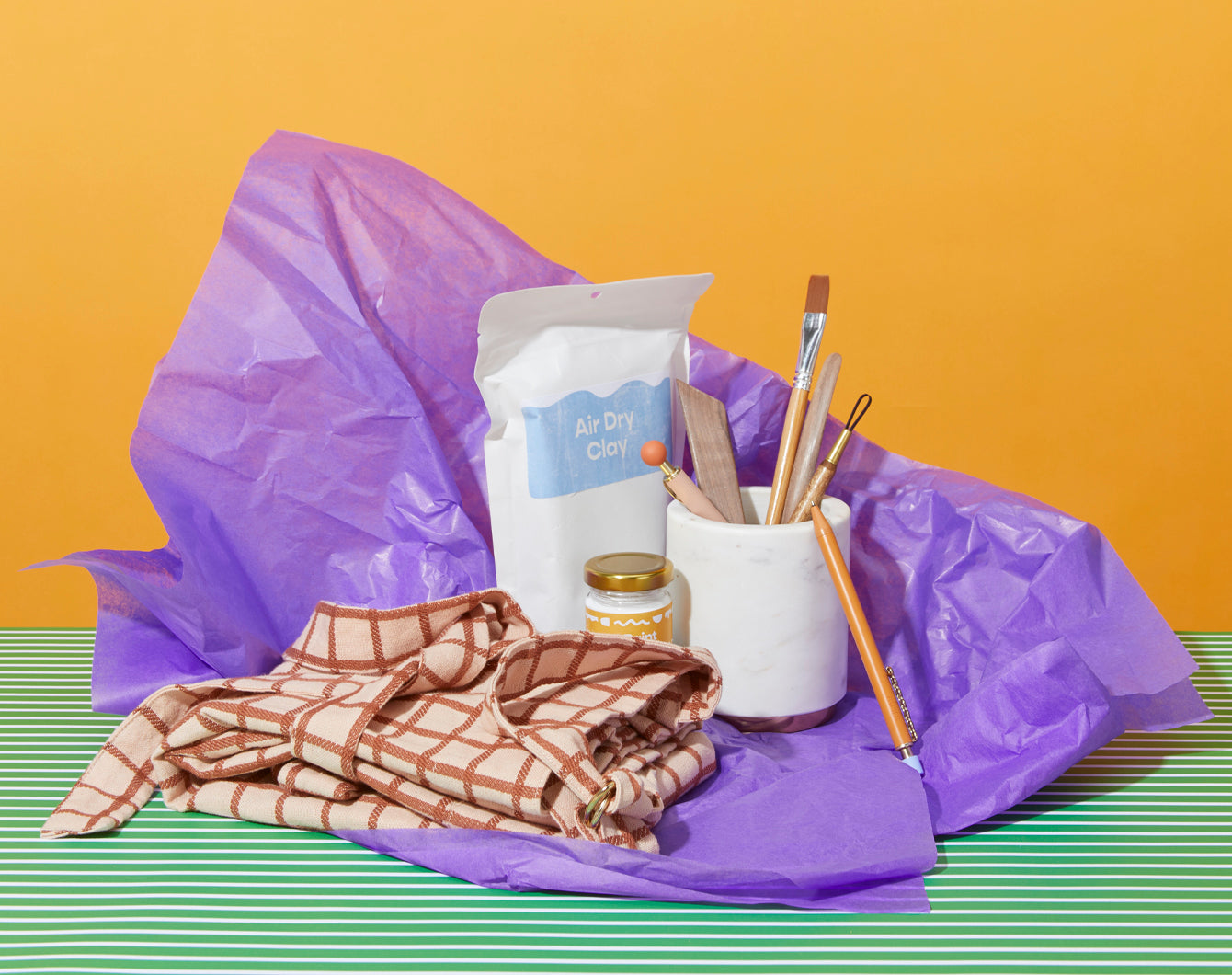A crafting set with air dry clay, a paintbrush, pencils, a cup, a jar, and a checked cloth on a striped surface with a yellow background.