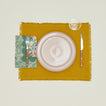 A table setting with blush plates, a clear glass, a mustard placemat, and a fern patterned napkin.