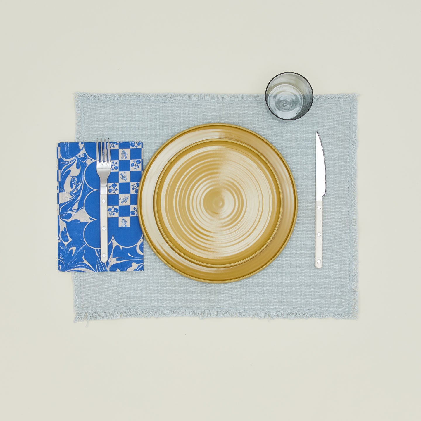 A table setting with mustard plates, a grey glass, a sky placemat, and a klein blue patterned napkin.